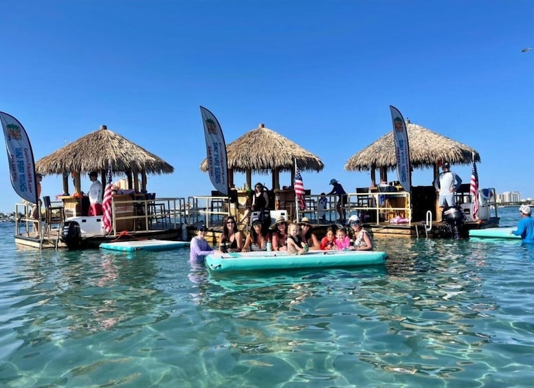 A group of people on a boat with thatched-roof structures, clear blue water, and flags. 1