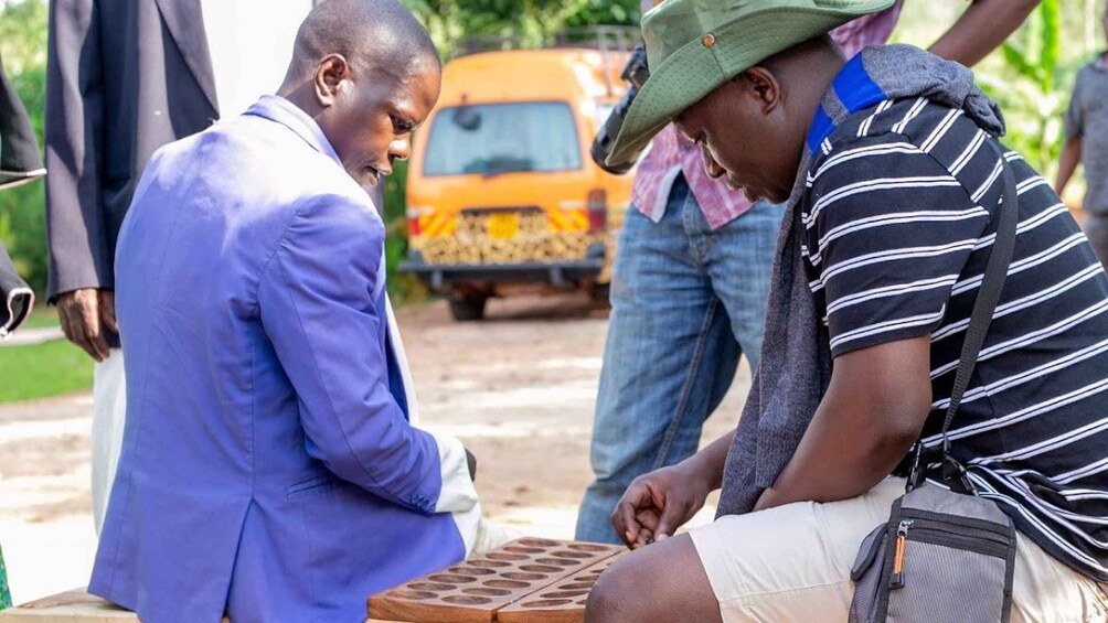 Two men engaged in a board game, with a vehicle and greenery in the background. 5