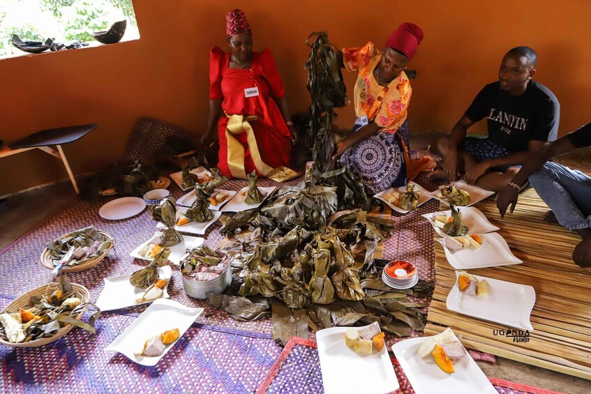 A group of people around a table with food wrapped in leaves, traditional attire, and a woven mat. 4