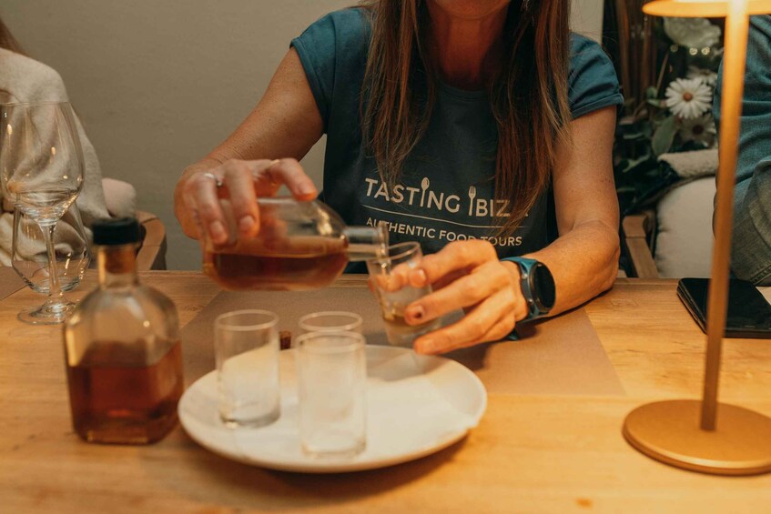 A person pouring a drink into a glass at a table with a bottle and shot glasses. 4