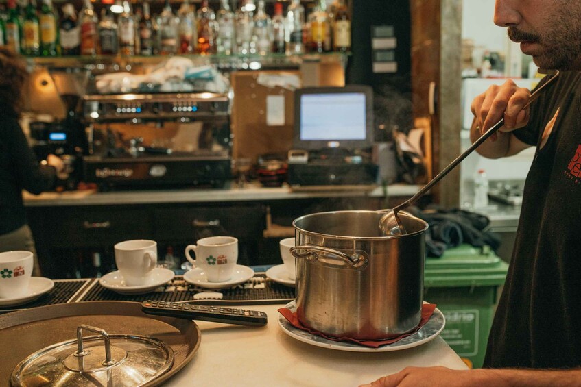 A barista stirring a pot on a tray with a spoon, with coffee cups and a computer monitor in the background. 3