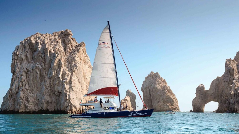 A sailboat with a red and white sail named 'Sail Rider' on clear turquoise waters, with rocky cliffs and a natural arch formation in the background. 3