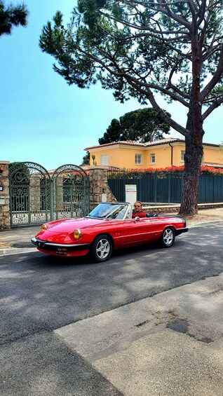 A red convertible car parked on a street with a stone gate, a tree, and a yellow building in the background. 3