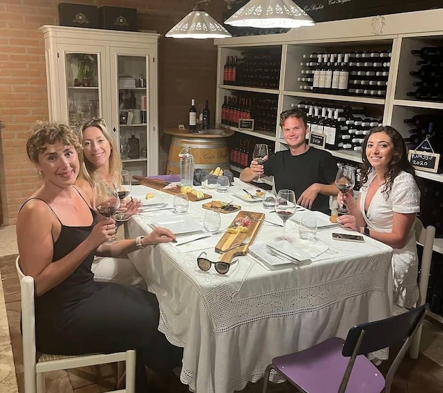 A group of people seated at a table with wine glasses, a wooden board with food, and wine bottles in the background. 1