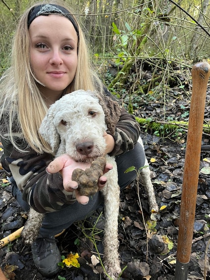 A person holding a dog in a natural setting with leaf-covered ground and a wooden post. 1