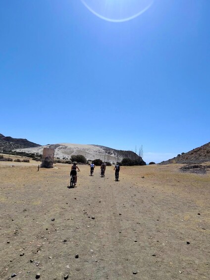 A group of cyclists on a dry, rocky terrain with a large rock formation in the background and a clear blue sky. 5