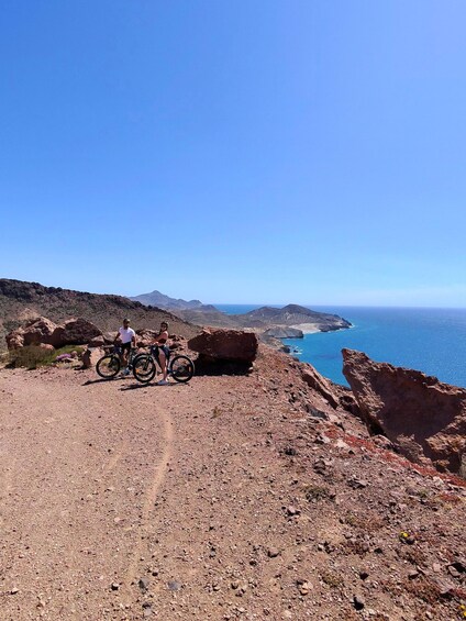 A rocky coastal landscape with two cyclists on a dirt path. 1