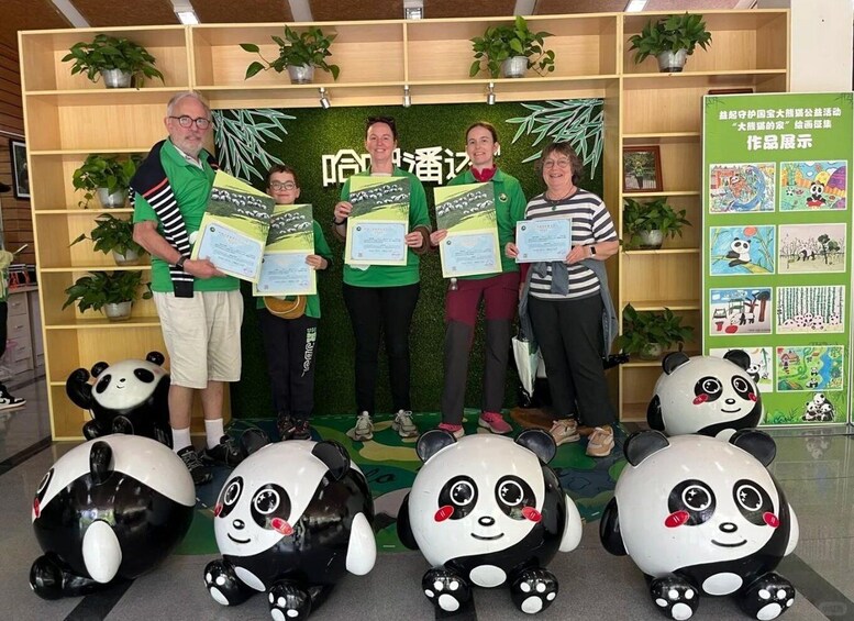 Four people holding certificates in front of a backdrop with panda illustrations and a panda sculpture. 1