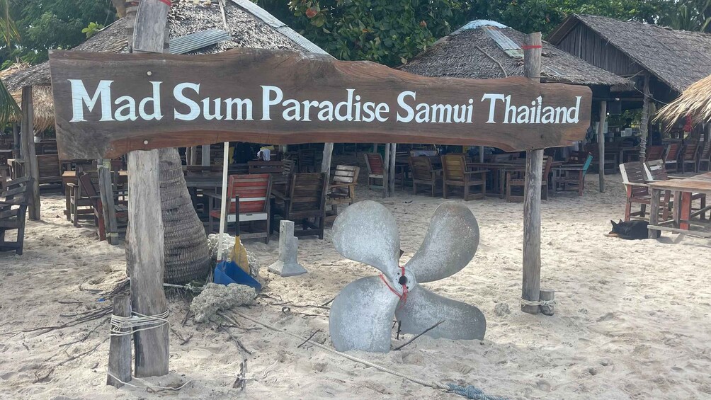 A wooden sign with 'Mad Sum Paradise Samui Thailand' written on it, surrounded by beach chairs and tables, with a thatched-roof structure in the background. 3