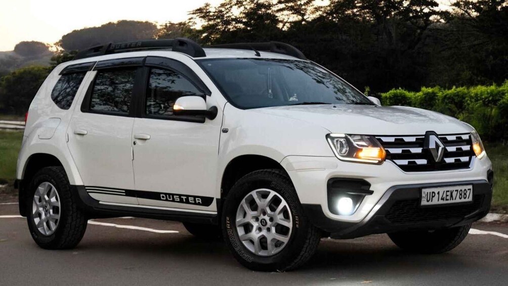 A white Duster SUV with a roof rack, on a road with greenery in the background. 5