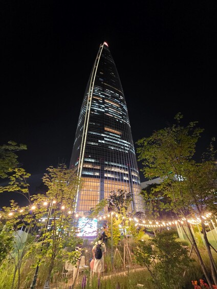 A tall, illuminated skyscraper at night with surrounding trees and string lights. 5