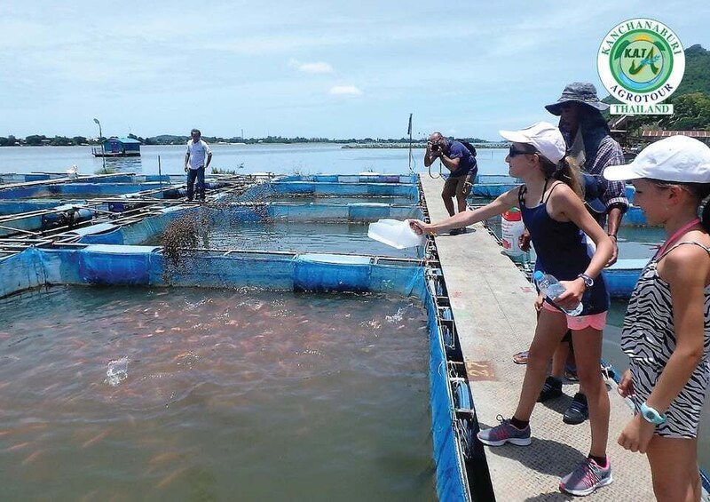 People on a dock near a body of water, some holding containers, with a sign for Kanchanaburi Agrotour Thailand in the background. 3