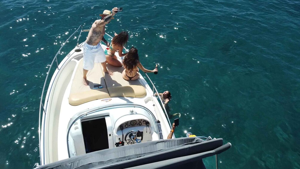 A boat with people on deck, some holding drinks, and a man swimming in the water. 4