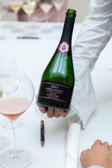 A person holding a bottle of 2015 Pierre Mignon champagne, a rose wine glass, and a pen on a white table. 2