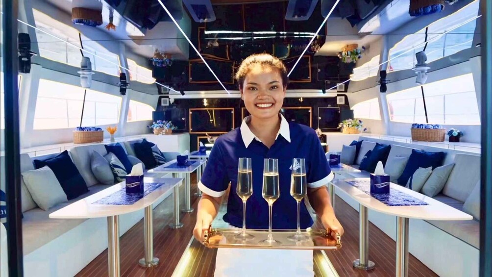 A person holding a tray with four champagne flutes in a modern, well-lit interior space with seating areas. 3