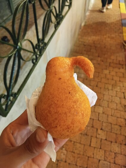 A hand holding a golden-brown fried food item with a paper wrapper, set against a brick pavement and a metal fence. 2