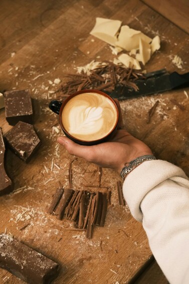 A hand holding a cup of coffee with latte art, surrounded by chocolate pieces and cocoa powder on a wooden surface. 1
