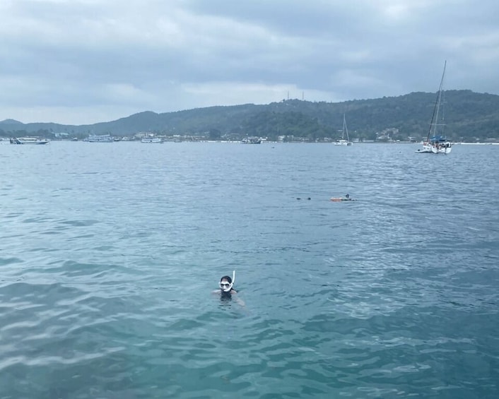 A calm sea with a person swimming, boats, and a hilly landscape in the background. 4