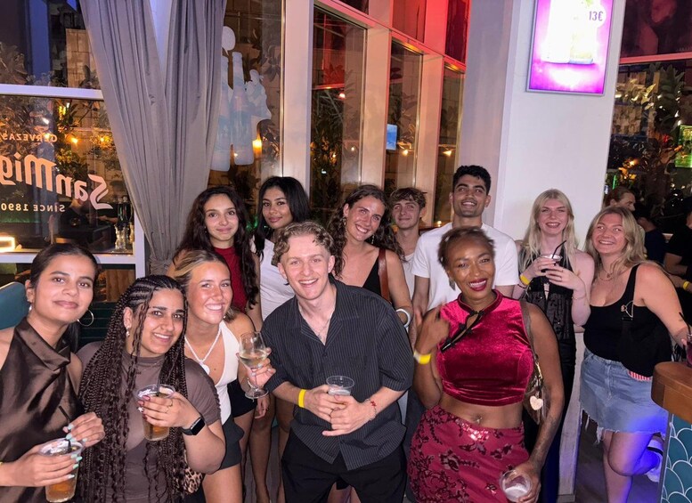 A group of people indoors, some holding drinks, with a bar and a window displaying a sign. 2