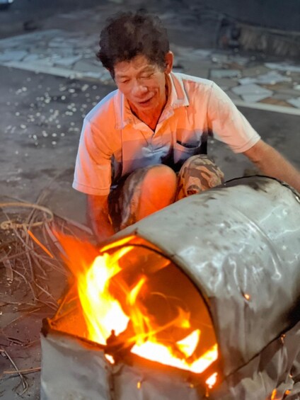 A person kneeling beside a fire burning inside a metal container. 1