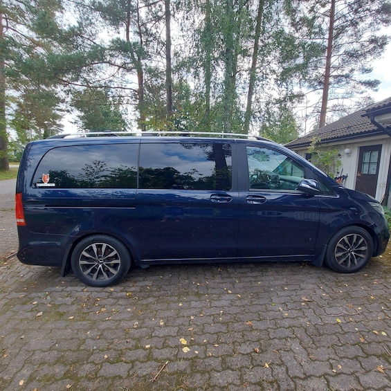 A dark blue minivan parked on a cobblestone driveway with trees and a small building in the background. 1