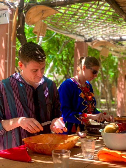 Two people preparing food outdoors with a wooden table, a woven basket, and a red cloth. 1