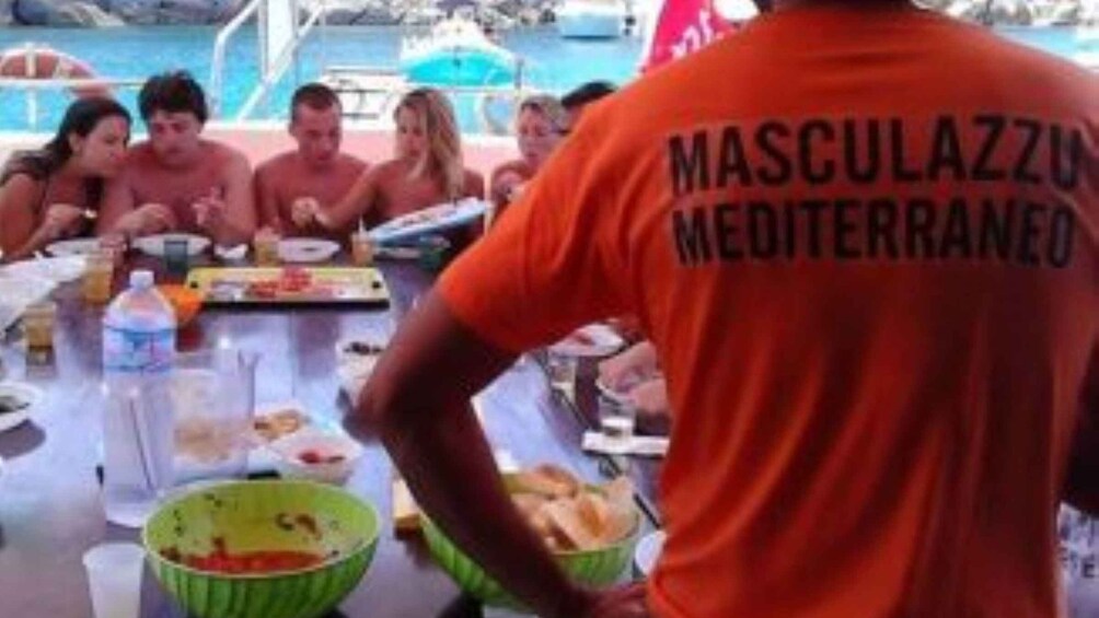 A group of people dining outdoors by a pool, with a person in an orange shirt labeled 'MASCULAZZU MEDITERRANEO' serving food. 5