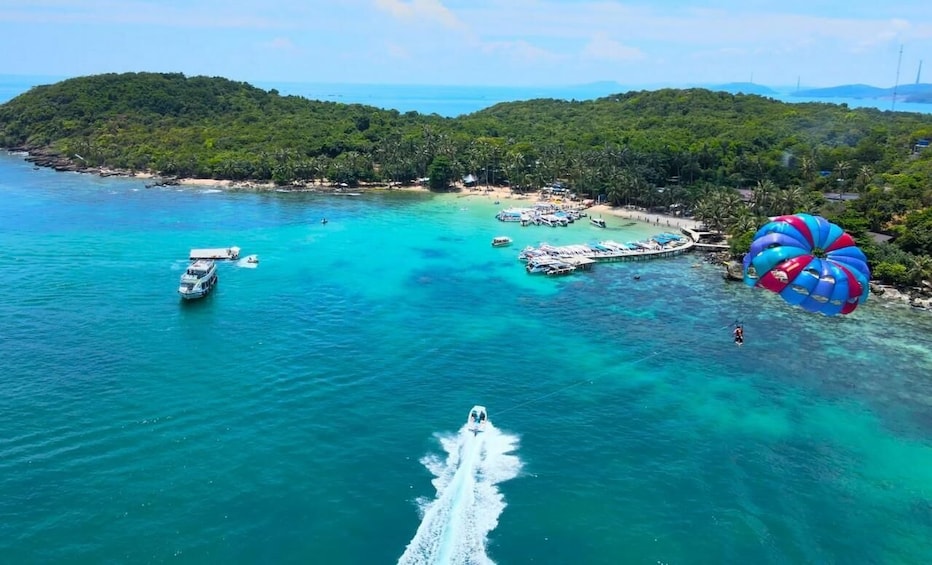 A clear blue sea with a boat speeding towards the shore, a parasail in the air, and a lush green island in the background. 2