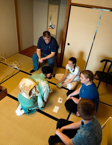 A group of people sitting on tatami mats in a room with a traditional Japanese aesthetic, including a shoji screen and a hanging scroll. 1