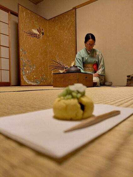 A person in traditional attire sitting at a low table with a bowl and chopsticks, in a room with a sliding door and a decorative panel. 3