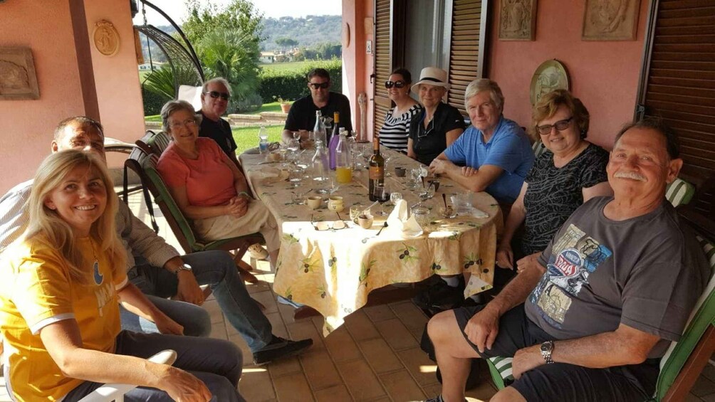 A group of people seated around a table with a tablecloth, in an outdoor setting with a view of greenery and buildings in the background. 3