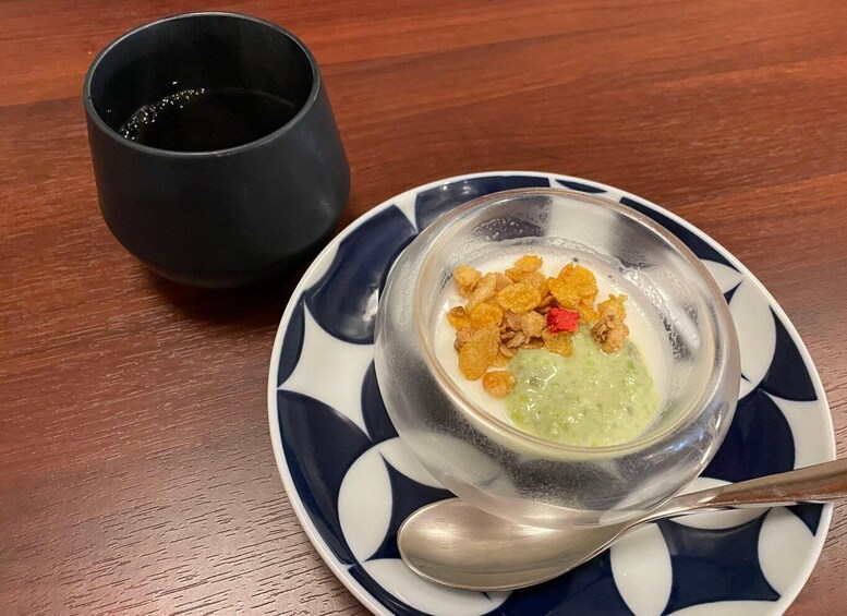 A bowl of soup with croutons and a spoon on a patterned plate, a black cup beside it, on a wooden table. 2