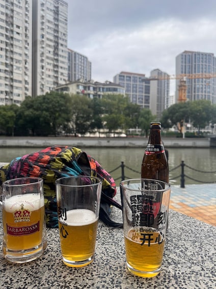Three glasses of beer, a bottle, and a colorful bag on a table by a waterfront with buildings in the background. 2