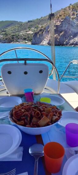 A table with a plate of pasta, a fork, a spoon, and a cup on a boat with a scenic view of a coastline and cliffs. 3