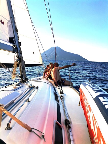 A sailboat with a person steering, a person lying down, and a mountainous landscape in the background. 1