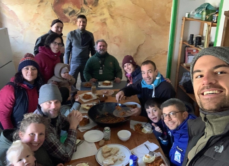 A group of people around a table with food, in a room with a textured wall and a green shelf. 2