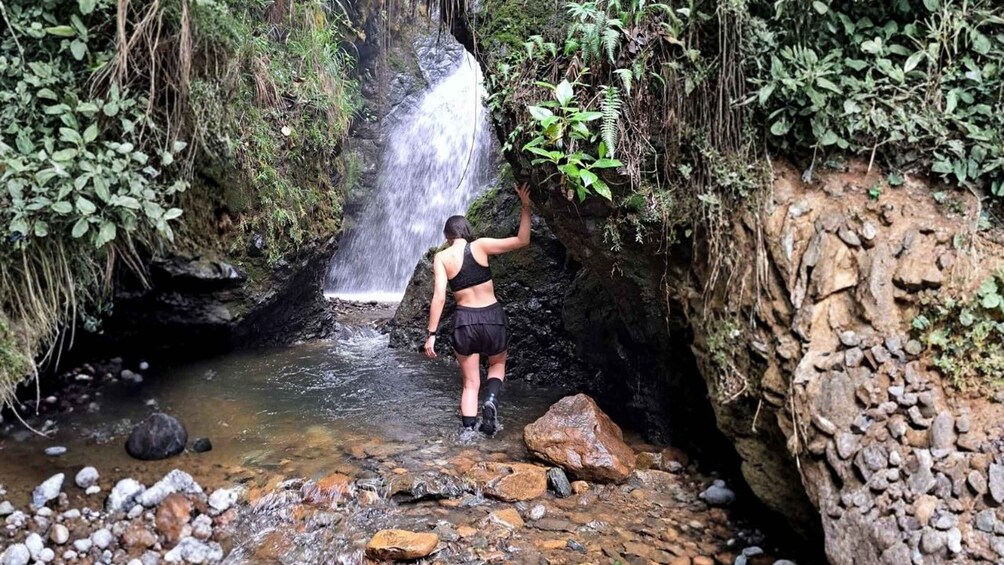 A person standing at the edge of a waterfall, surrounded by rocks and greenery. 1