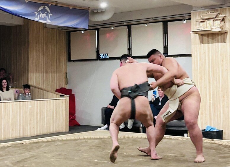 Two sumo wrestlers engaged in a match, with spectators in the background. 1