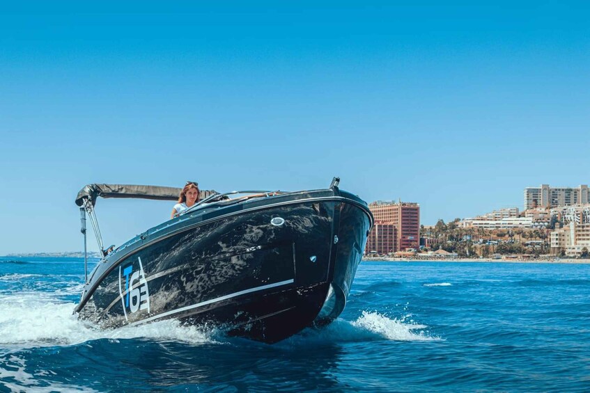A black speedboat with a person on board, moving through clear blue water with a cityscape in the background. 1