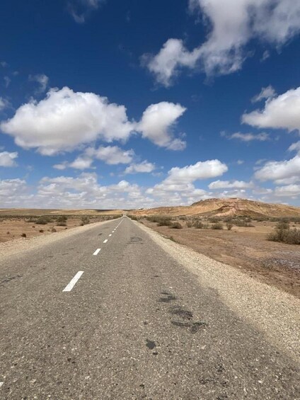 A road stretching through a desert landscape with sparse vegetation and distant hills under a partly cloudy sky. 3
