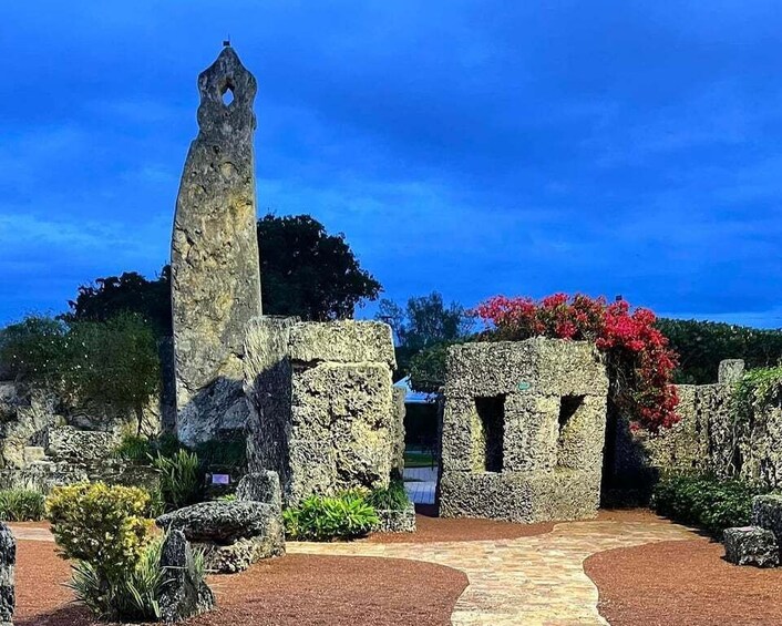 A stone monument with a pointed top, surrounded by a garden with red flowers and a paved walkway. 4