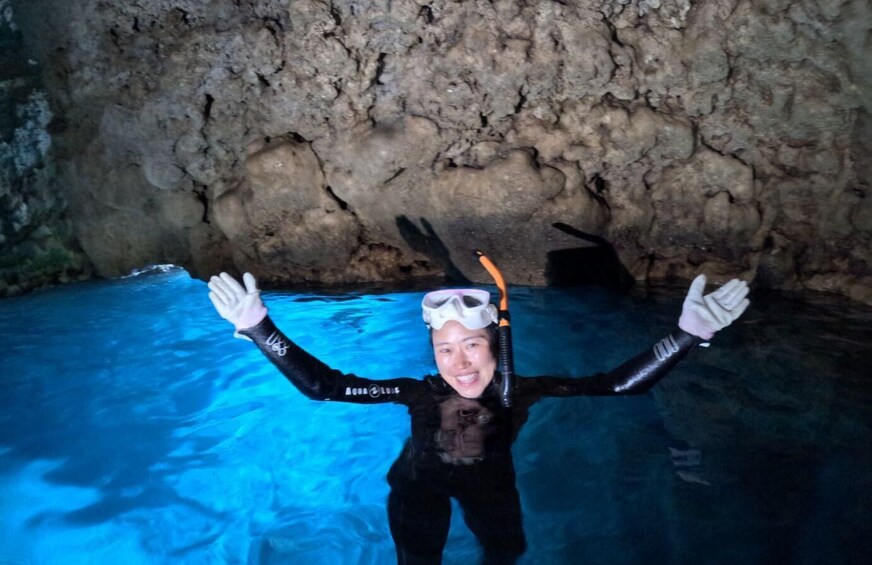 A person in a black wetsuit with arms raised, standing in a pool with a rocky cave wall in the background. 2