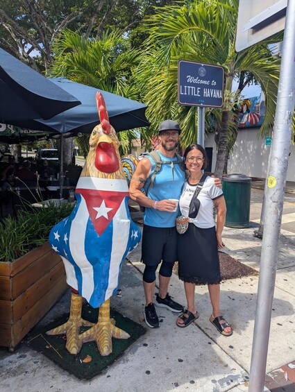 A person in a costume resembling the Cuban flag, a sign reading 'Welcome to Little Havana, USA,' and a street scene with palm trees and outdoor seating. 1
