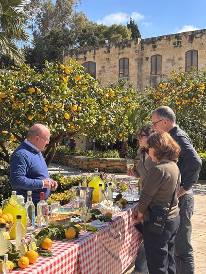 A group of people at an outdoor table with a checkered tablecloth, surrounded by lemon trees and a stone building in the background. 1