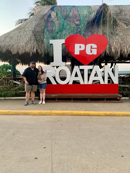 A couple standing in front of a sign that says 'I ♥ PG ROATAN' with a thatched roof structure behind them. 4