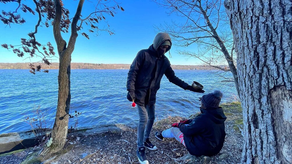 A person standing by a lake, holding a metal bowl, with another person sitting nearby. 1
