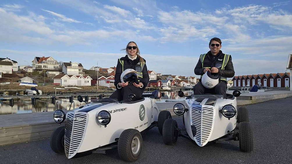 Two people on small white vehicles with 'HOT ROD RIDERZ' branding, parked by a waterfront with houses in the background. 1