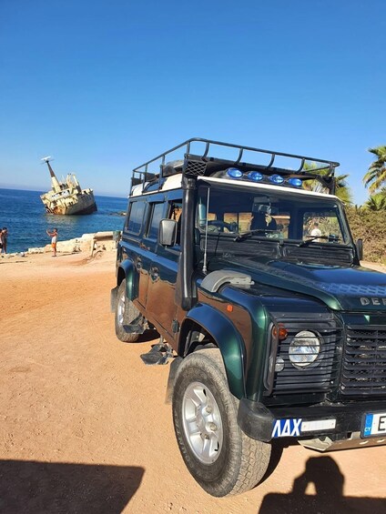 A dark green off-road vehicle parked on a sandy beach with a shipwreck in the background. 1