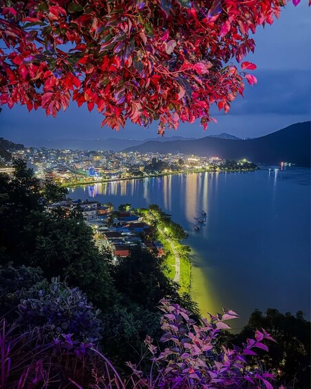 A nighttime cityscape with a river reflecting lights, surrounded by hills and dense foliage. 2