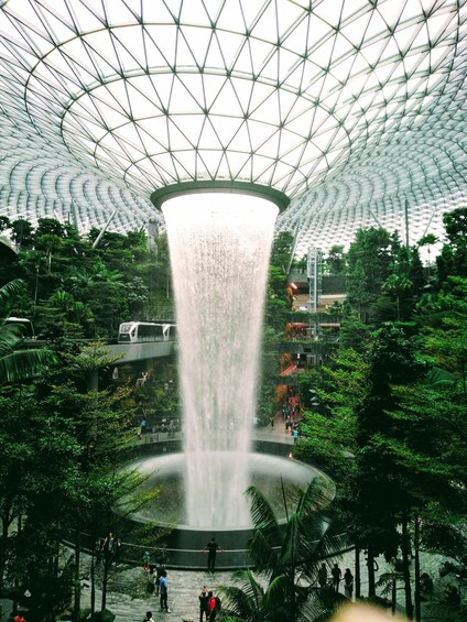 A large indoor waterfall with a glass ceiling, surrounded by lush greenery and people walking around. 1